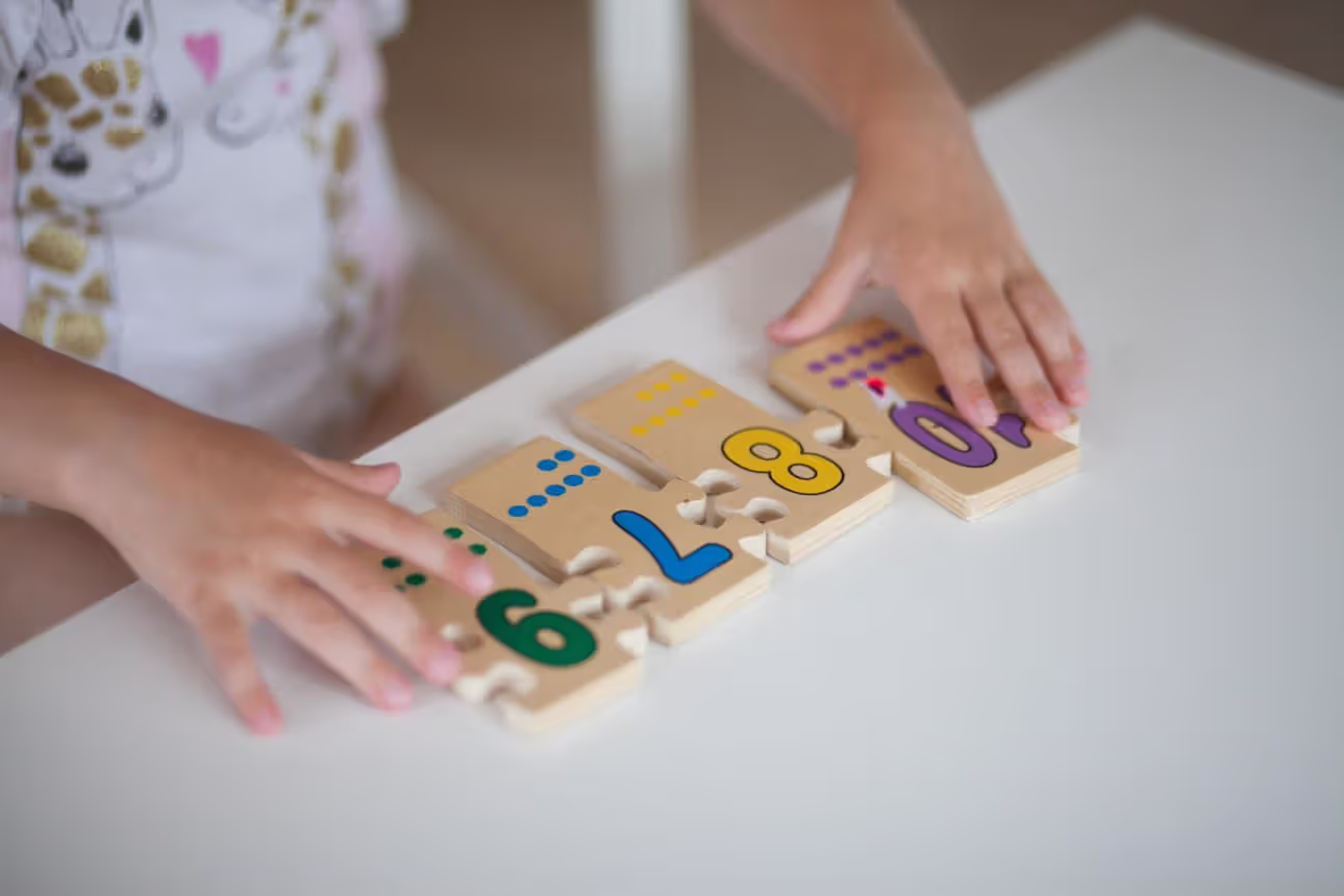 Реалистичная сцена: ребёнок preschool возраста сидит за столом с цветными pattern blocks и карточками последовательностей; ребёнок пытается повторить простой цветовой паттерн, воспитатель мягко показывает пример; акцент на pattern recognition difficulties; modern preschool classroom, photorealistic, high detail, soft daylight, 16:9, no readable text, no logos, compressed strictly under 1MB.